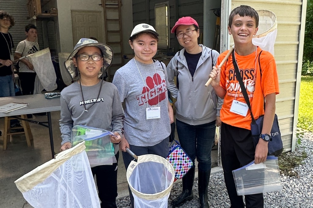 Children wearing hiking clothes and holding butterfly nets, smile for a group photo outside.