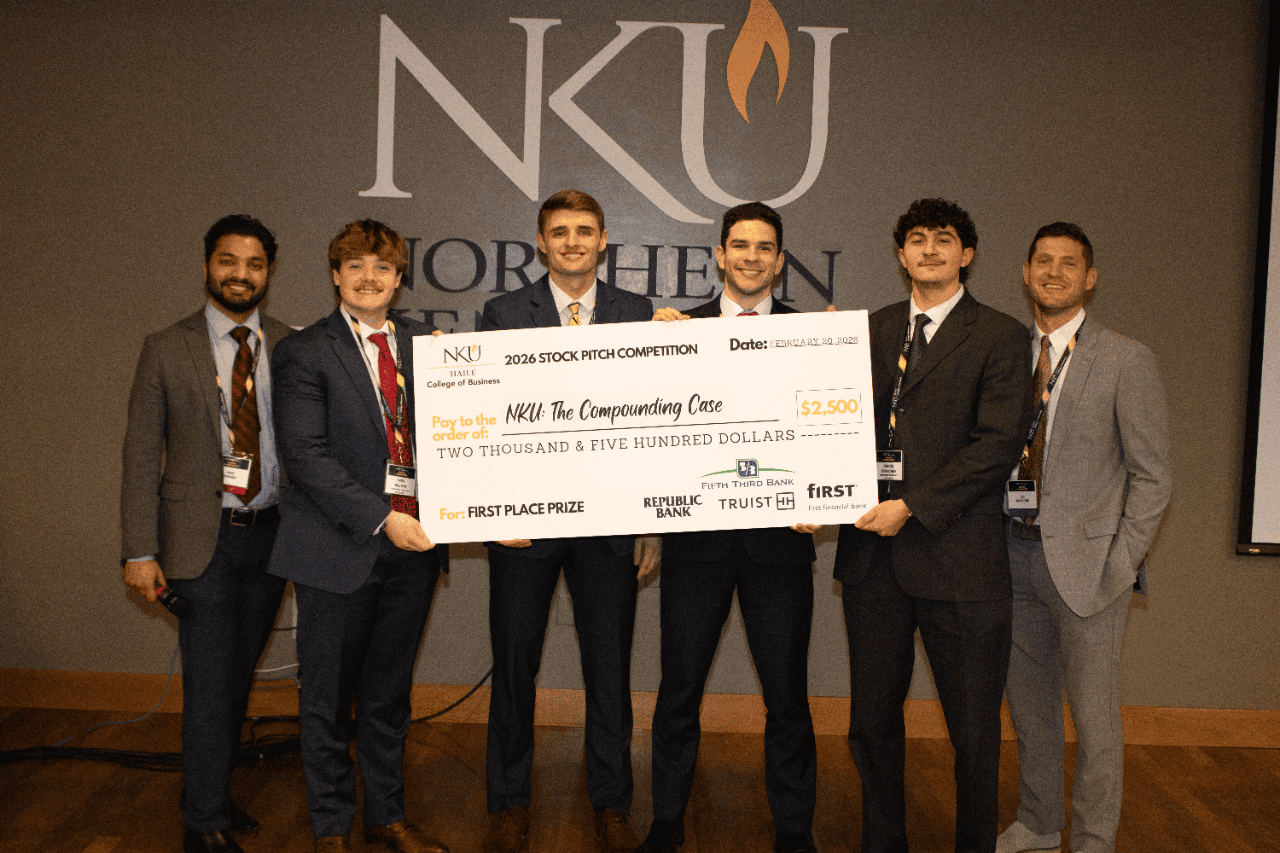 Four male students pose with a giant check and two faculty members in front of the NKU logo.